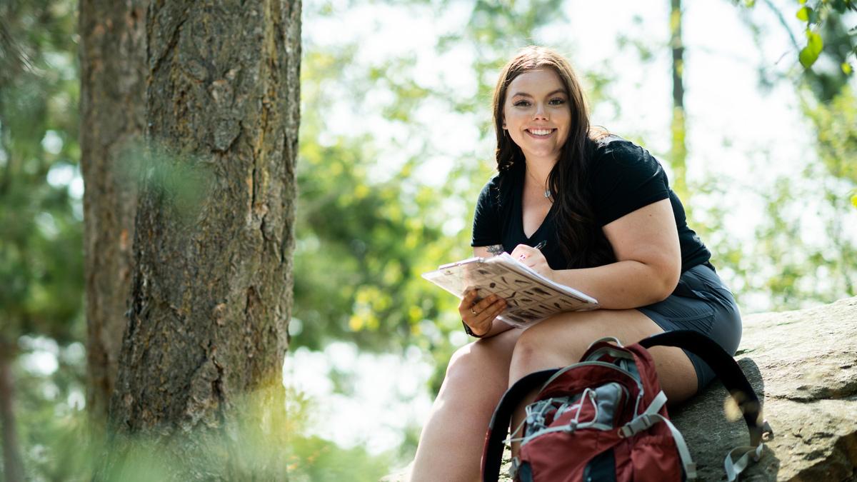 Emma Arman looks up from her journaling and smiles while sitting on a boulder among the trees along Tubbs Hill in Coeur d’Alene.